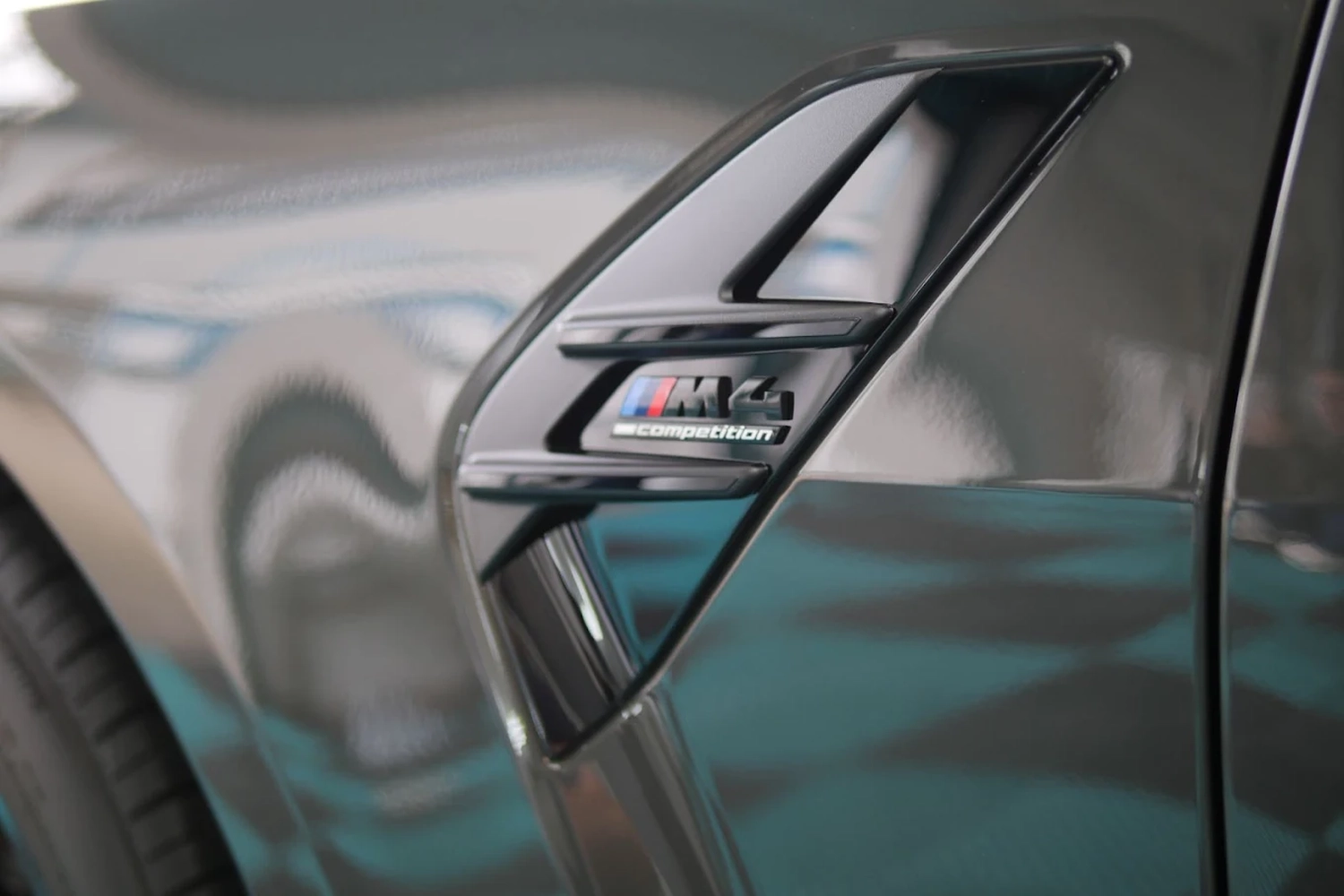 Simons Garage - Fahrzeugaufbereitung - Nahaufnahme eines glänzend grauen BMW M4 Competition-Emblems an der Seite.