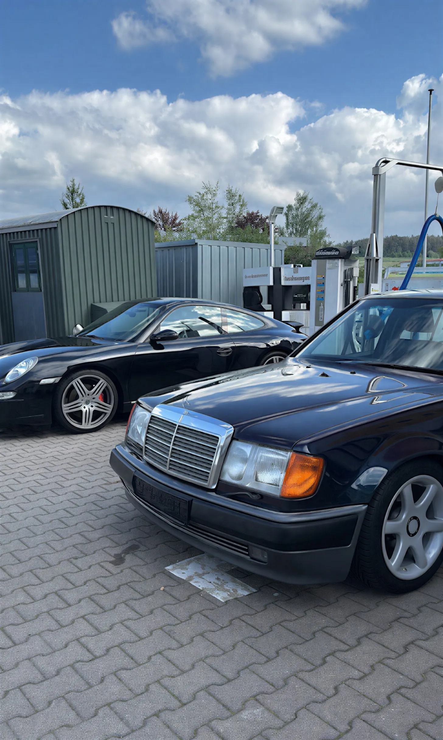 Simons Garage - Fahrzeugaufbereitung - Eine dunkelblaue Mercedes-Benz W124 Limousine, geparkt neben einem schwarzen Porsche 911 Coupé im Freien.