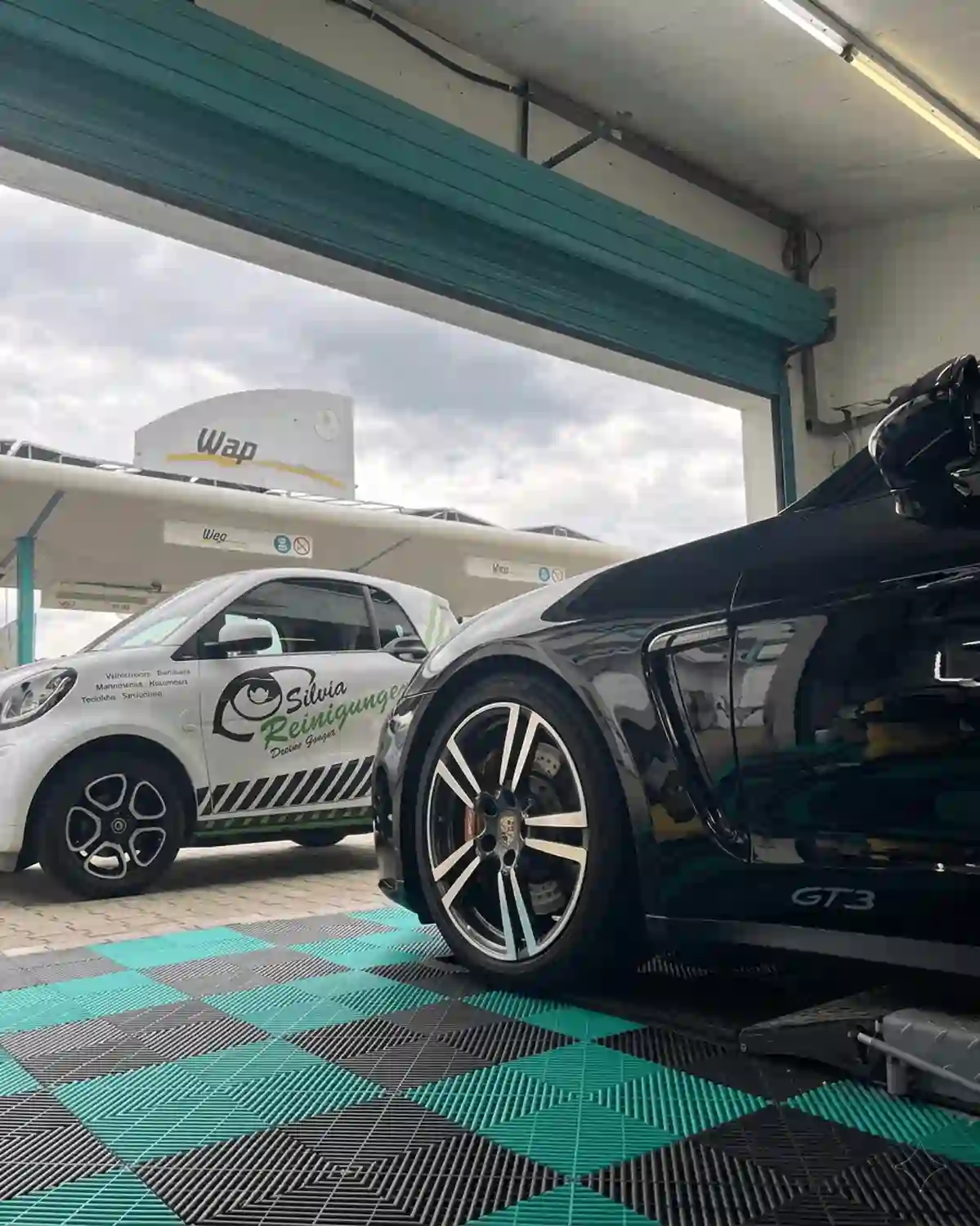 Simons Garage - Fahrzeugaufbereitung - Seitenansicht eines schwarzen Sportwagens vor einer Waschanlage neben einem weißen Servicefahrzeug.