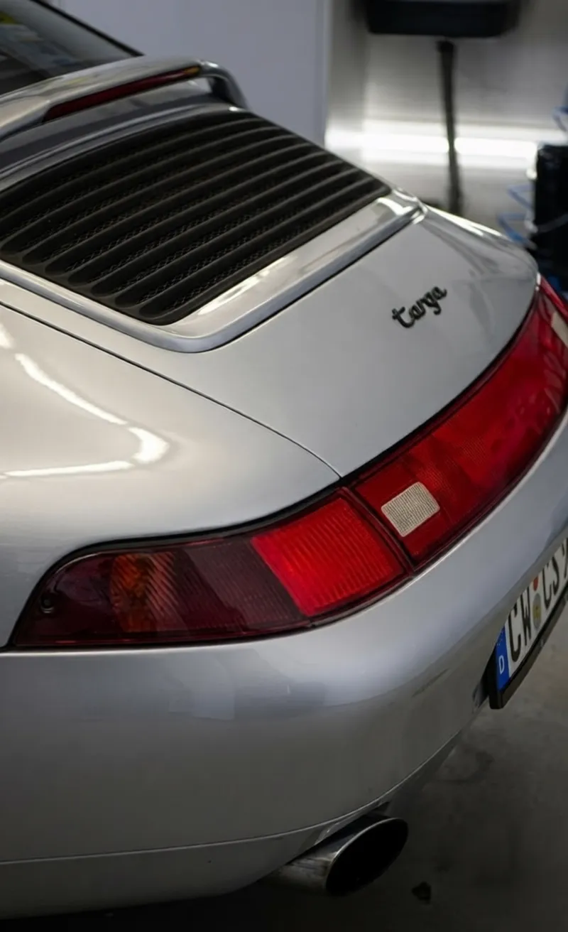 Simons Garage - Fahrzeugaufbereitung - Nahaufnahme der hinteren Motorlüftungsschlitze und der Rückleuchtenleiste mit „Targa“-Schriftzug eines silbernen Porsche.