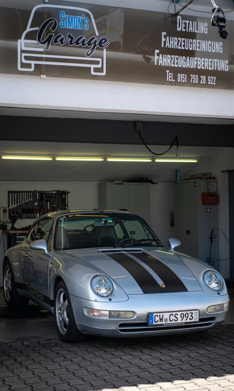 Simons Garage - Fahrzeugaufbereitung - Frontansicht eines klassischen silbernen Porsche-Sportwagens mit schwarzen Rennstreifen in einer Garage.