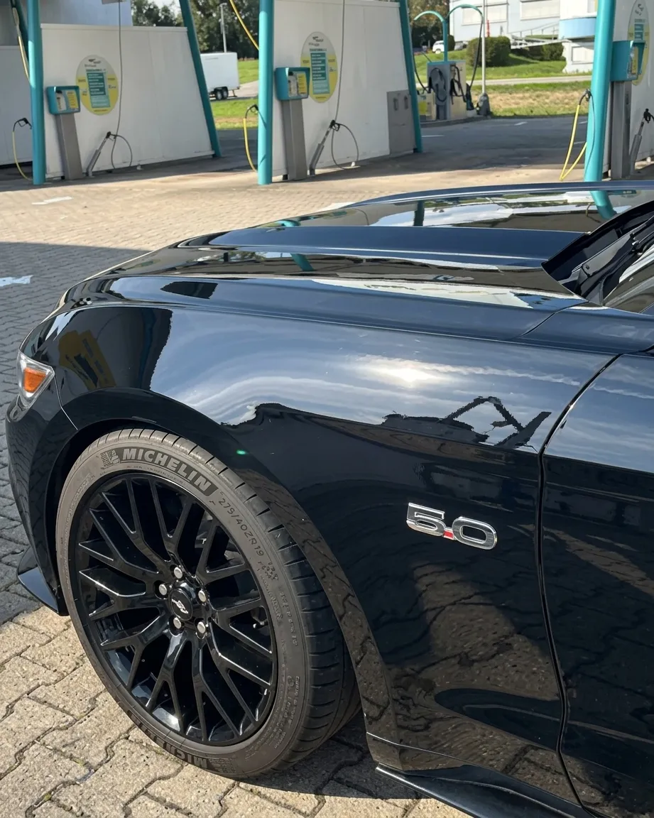 Simons Garage - Fahrzeugaufbereitung - Seitenansicht eines glänzend schwarzen Sportwagens mit schwarzem Michelin-Reifen und „5.0“-Emblem.