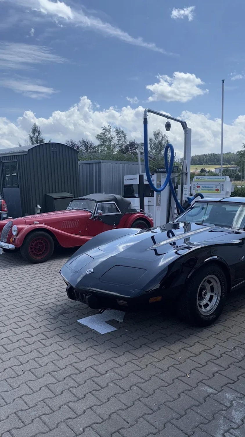 Simons Garage - Fahrzeugaufbereitung - Glänzend schwarze Chevrolet Corvette im klassischen Stil, geparkt im Freien neben einem roten Oldtimer-Roadster.